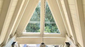 Een kamer met hoge plafonds en een schuine hemel, een hanglamp, een zithoek met tafel en stoelen, en uitzicht op groen buiten.