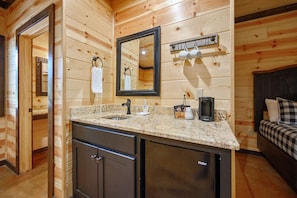 Compact bathroom with a modern design, featuring a granite countertop, sink, and mirror, as well as a coffee maker and dark-toned cabinet.