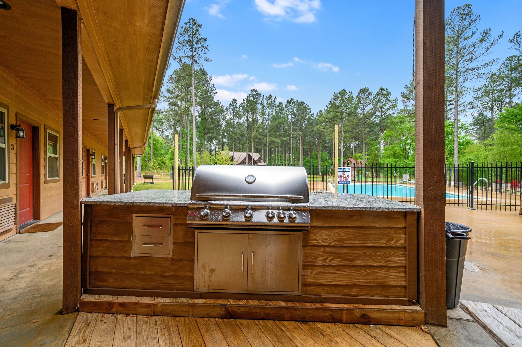 Cucina esterna con barbecue, terrazza in legno e vista su piscina e alberi.