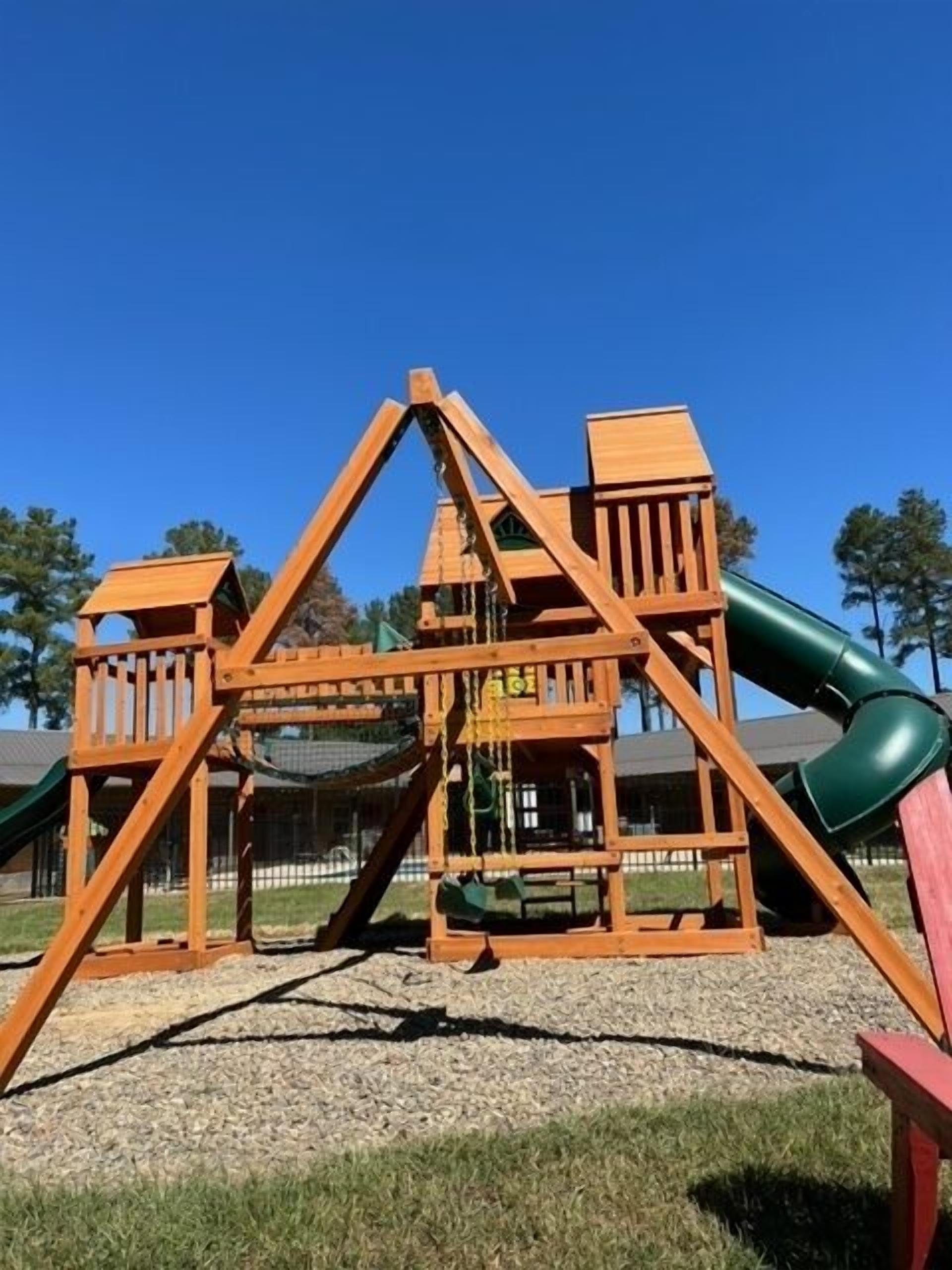 A wooden playground structure with a slide and climbing elements, set on a gravel surface with grass around, under a clear blue sky.