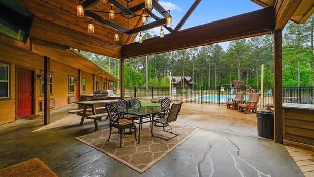 A covered patio with a table and chairs, a pool, and a view of the surrounding forest.