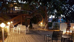 Une terrasse extérieure dotée d’un plancher en bois, de chaises et de tables en métal blanc, entourée d’arbres et d’une clôture en bois.