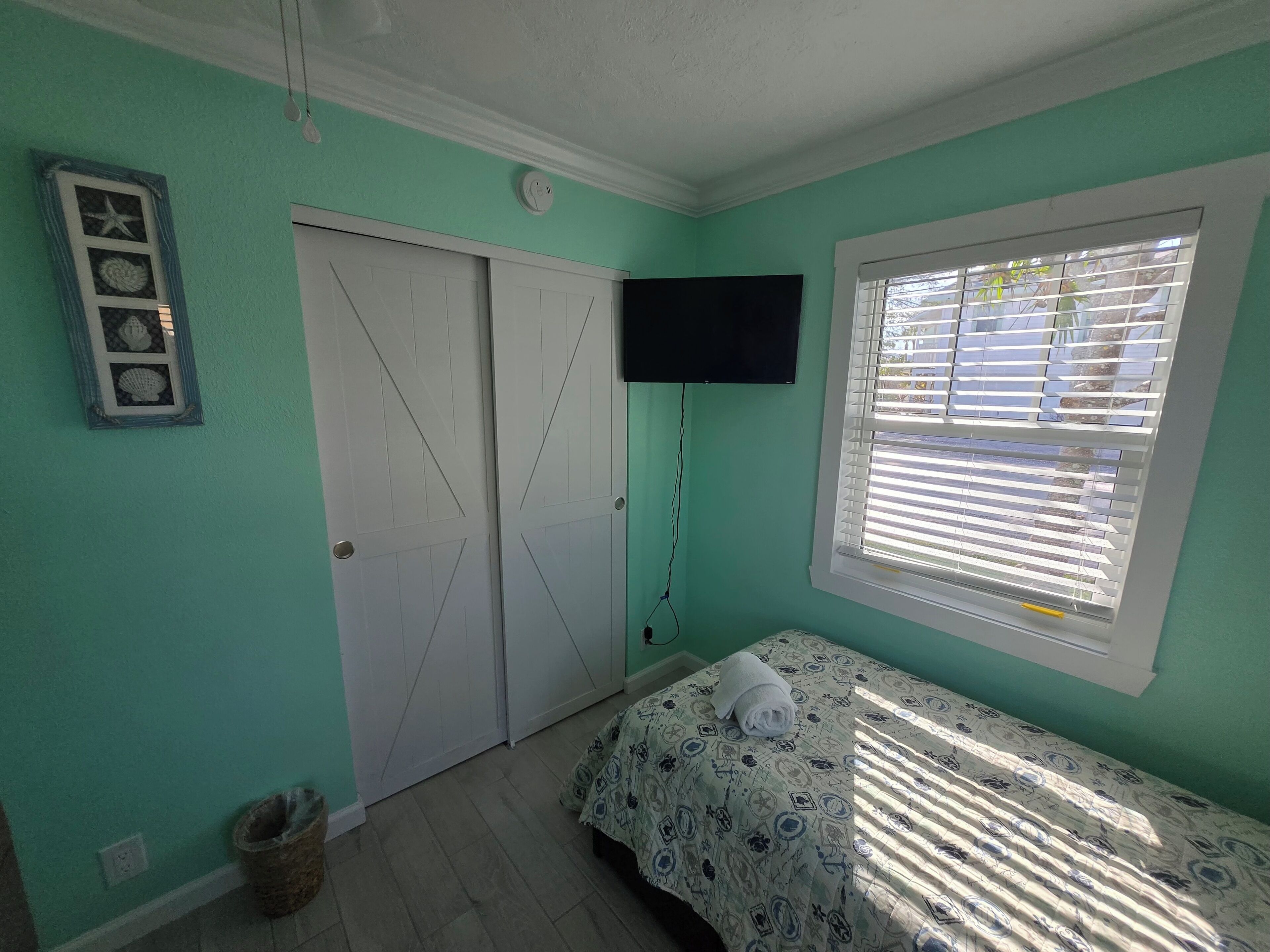 Une pièce aux murs de couleur turquoise, un lit recouvert d’une couvre-lit à motifs, un téléviseur fixé au mur, une fenêtre avec des stores et une porte décorée d’une étoile.