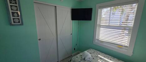 Une pièce aux murs de couleur turquoise, un lit recouvert d’une couvre-lit à motifs, un téléviseur fixé au mur, une fenêtre avec des stores et une porte décorée d’une étoile.