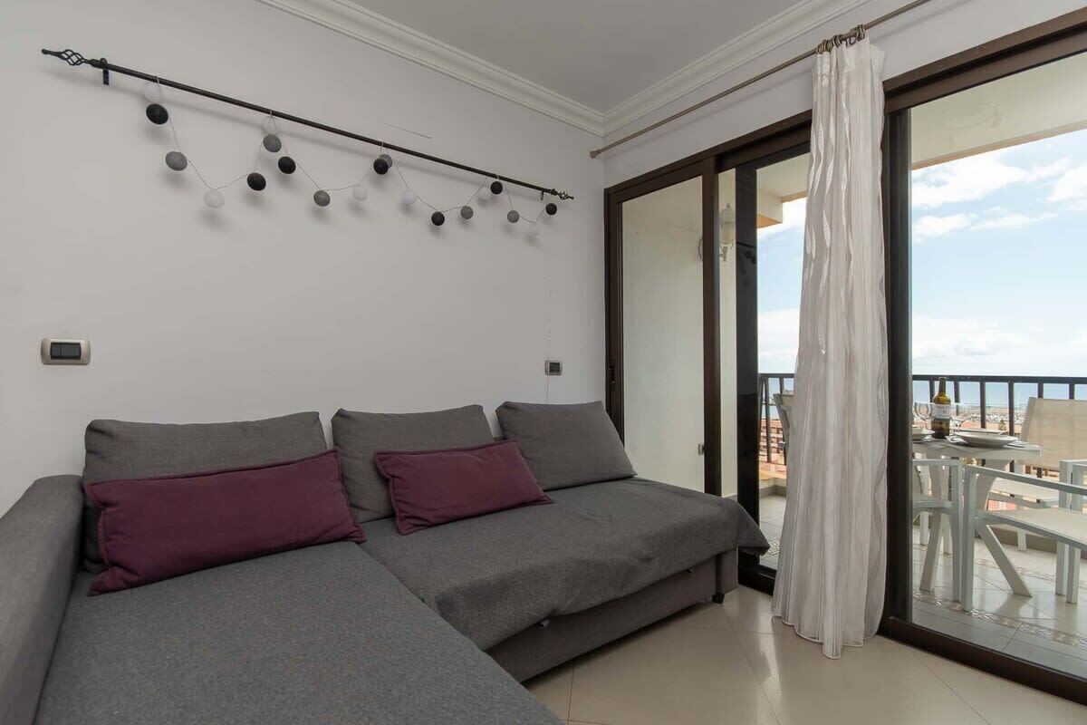 A modern living room with a grey sofa set, a string of lights on the wall, and a balcony with outdoor furniture.