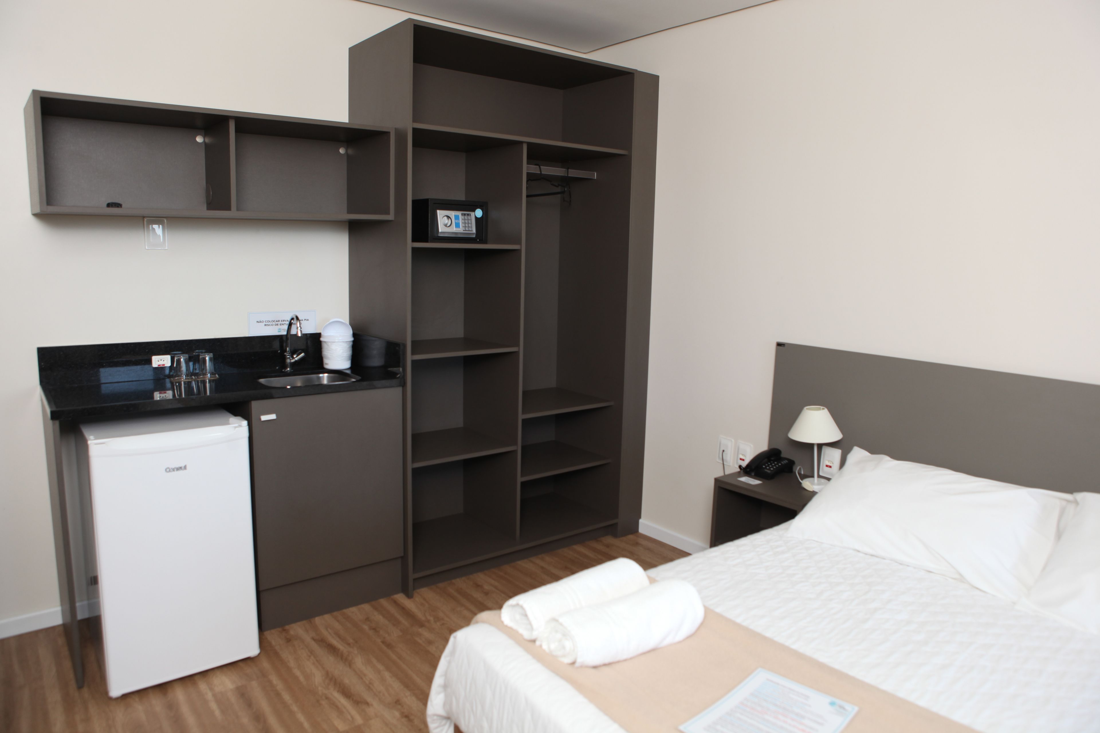 Une chambre d’hôtel compacte comprenant un lit, une petite kitchenette, une armoire intégrée et une table de chevet avec une lampe.