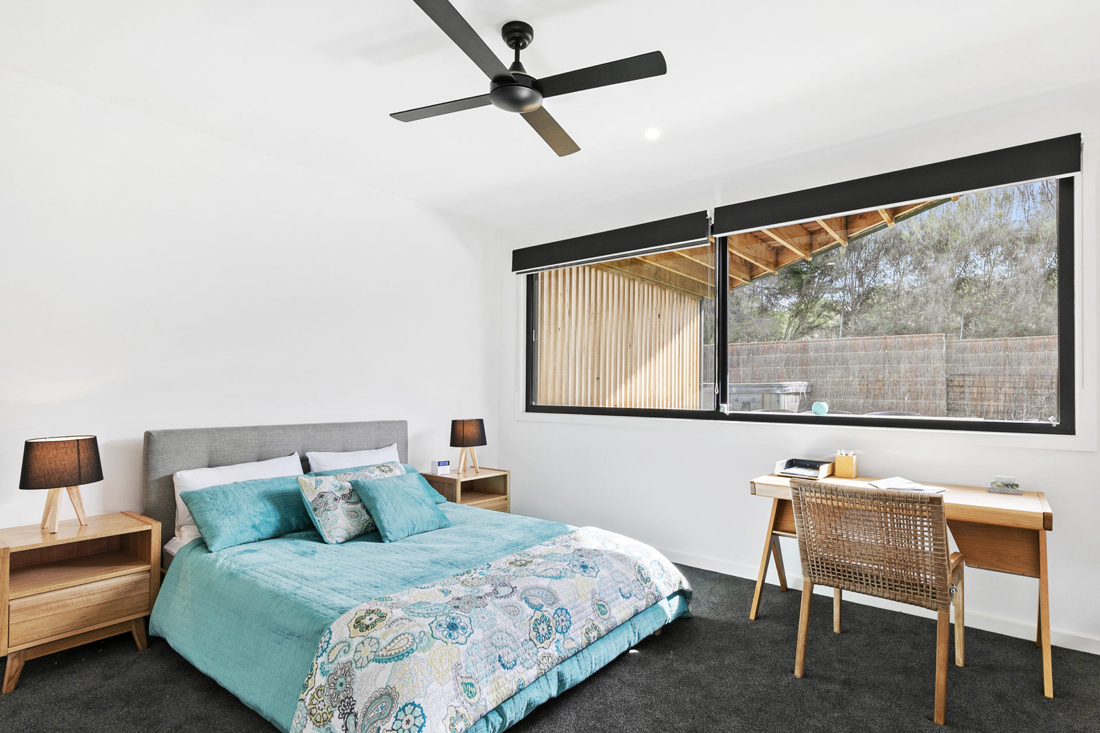 A bedroom with a bed, two bedside tables, a desk, a chair, a ceiling fan, and a large window.
