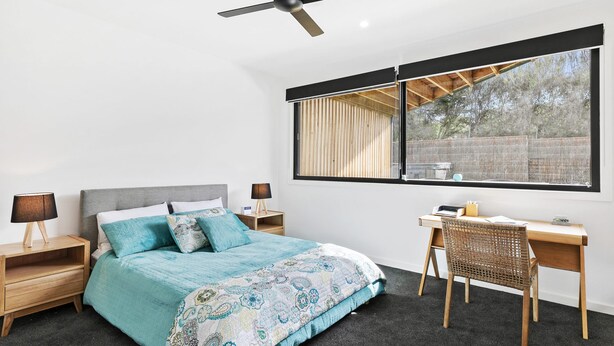 A bedroom with a bed, two bedside tables, a desk, a chair, a ceiling fan, and a large window. - Point Break (Anglesea)