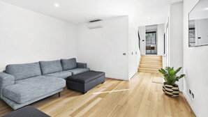 A modern living room with a grey sectional sofa, a dark ottoman, wooden flooring, a potted plant, and a staircase leading to another room. - Point Break (Anglesea)