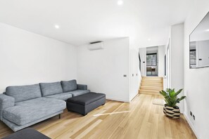 Ein modernes Wohnzimmer mit einem grauen Ecksofa, einem dunklen Hocker, Holzböden, einer Topfpflanze und einer Treppe, die in einen anderen Raum führt.