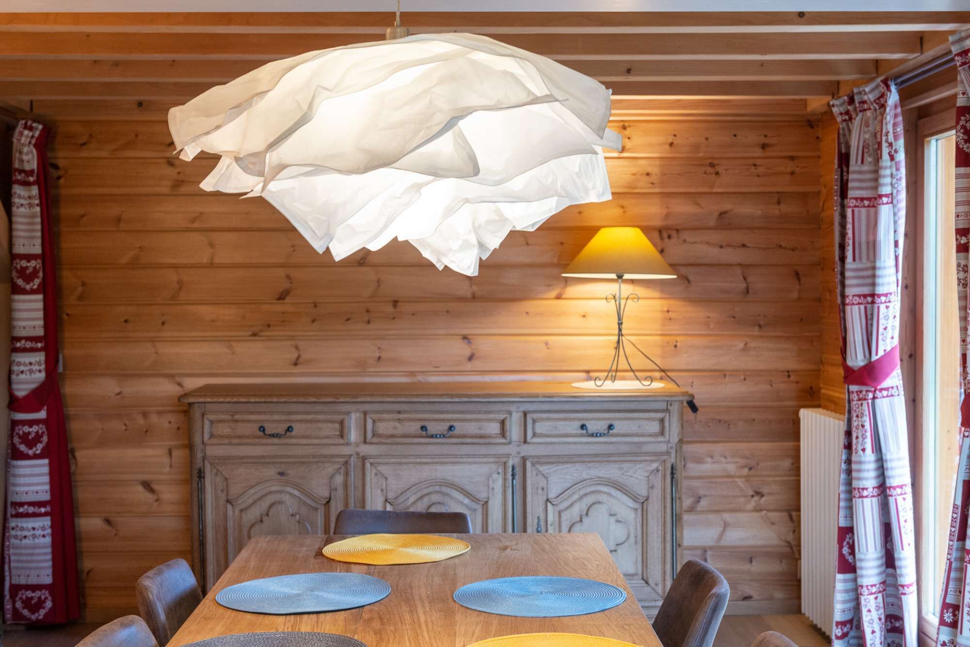 Ein Holzblockhaus-Interieur mit einem Esstisch, Stühlen, einer Lampe und einem gemusterten Vorhang.