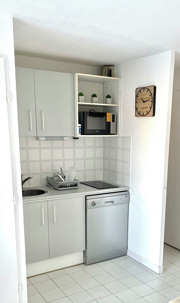 Une cuisine compacte avec des meubles blancs, un four à micro-ondes, un lave-vaisselle et une horloge murale.