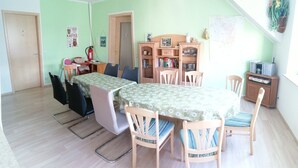 Un comedor con una mesa dispuesta para cuatro personas, rodeada de sillas. Hay una estantería con libros y objetos decorativos, un mueble bajo con un televisor, y un mapa en la pared.