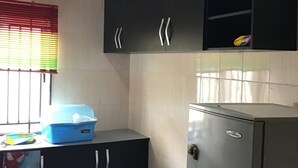 A compact kitchen with black cabinets, a stainless steel refrigerator, and a window with blinds. - Gees Shortlets Apartments (Lagos)