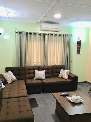 A living room with a sectional sofa, a coffee table, and a wall-mounted air conditioner. - Gees Shortlets Apartments (Lagos)