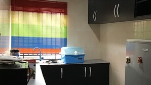 A kitchen with black cabinets, a refrigerator, a window with colorful blinds, and a countertop with a blue container. - Gees Shortlets Apartments (Lagos)