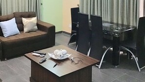 A living room with a brown sofa, a glass coffee table, and a dining table with chairs. - Gees Shortlets Apartments (Lagos)