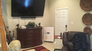 A living room with a flat-screen TV mounted on the wall, a wooden cabinet, a white chair, a brown leather chair, and a whiteboard with writing on it. - Professional, Travelers Haven , 1 bdrm, living room, private shower free parking (Martinez)