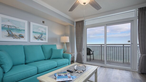 A teal sofa, a wooden coffee table with magazines and a remote, and a balcony with a view of the ocean.