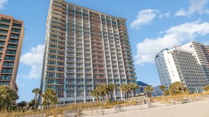 A high-rise hotel with multiple balconies, located near a beach with a sandy foreground and palm trees.