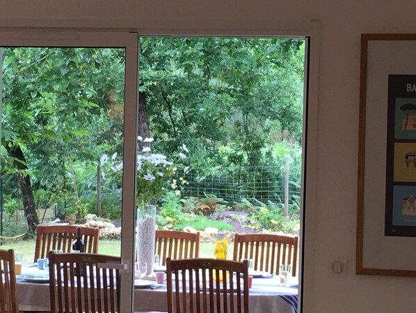 Un comedor interior con sillas y mesas de madera, una puerta de cristal que da a un jardín con árboles y una valla.