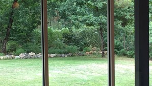 Ein Zimmer mit großen Fenstern und Blick auf einen Rasen mit Bäumen und einer Steinmauer.