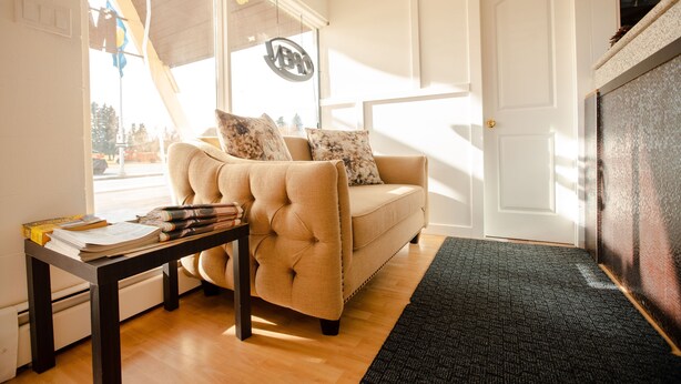 A living room with a beige sofa, a small table with books, and a black rug. - Camrose Motel (Camrose)