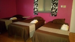 A room with three single beds, each with a pillow and a towel. The walls are pink, and there are two windows with floral curtains. - Labrish Guest House (Kingston)