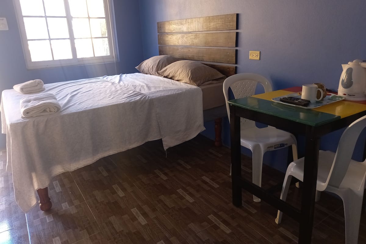 A room with a bed, a table with a kettle, a chair, and a window.