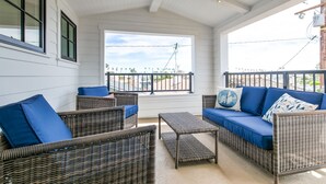 A covered outdoor seating area with wicker furniture and blue cushions, large windows with a view of the street, and a white exterior wall.