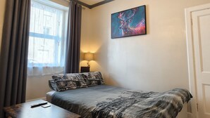 Une chambre à coucher comprenant un lit, un bureau, une lampe, une fenêtre avec des rideaux et un tableau accroché au mur.