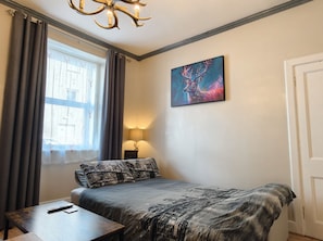 Een slaapkamer met een bed, een bureau, een lamp, een raam met gordijnen en een schilderij aan de muur.
