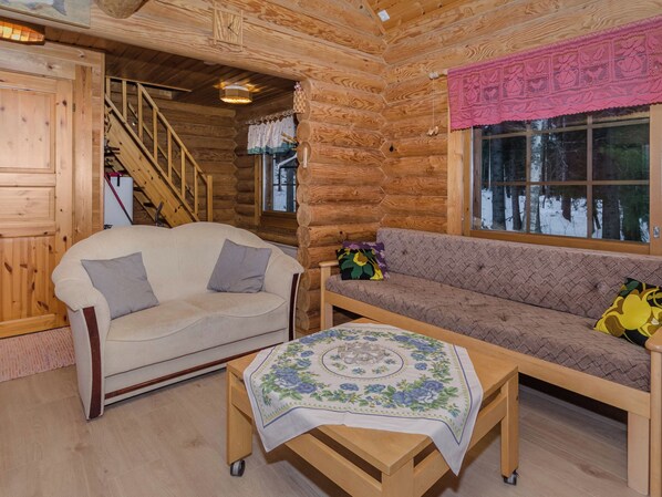 A log cabin interior with a sofa, a chair, a table with a tablecloth, a staircase, and a window with a view of trees.