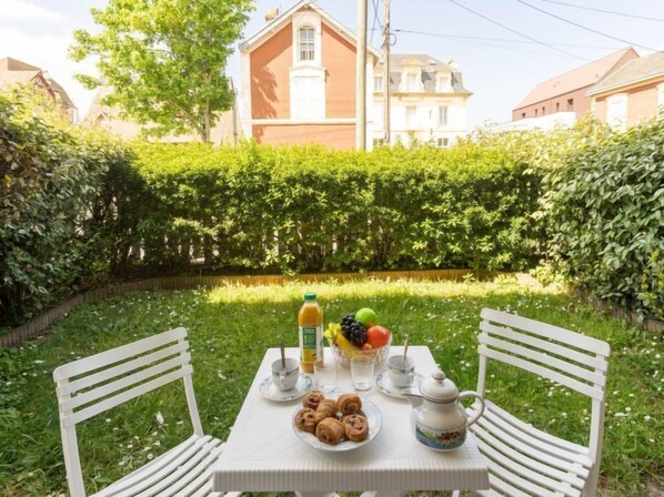 An outdoor dining table with a breakfast setting, including croissants and fruit, in a garden with a hedge and buildings in the background.