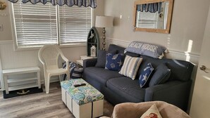 A living room with a dark grey sofa, a white chair, a patterned ottoman, and a beige armchair. There is a window with blinds and a mirror on the wall. - Beach Block Wildwood Crest Condo (Wildwood Crest)