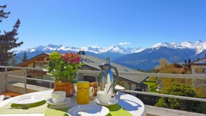 A breakfast setup on a balcony with a mountain view, including a table with plates, cups, a pitcher, and a vase with flowers. - Jeanne D'Arc Apt. E-38 by Interhome (Crans-Montana)
