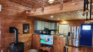 A cozy cabin interior with a wood stove, kitchen area, and a TV on a wooden stand. - Beautiful and modern cape style cottage nested along the Jordan Tidal River. (Lamoine)