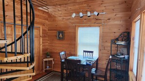 A wooden cabin interior with a dining table, chairs, a small side table, a window with blinds, a spiral staircase, and a shelf with items. - Beautiful and modern cape style cottage nested along the Jordan Tidal River. (Lamoine)