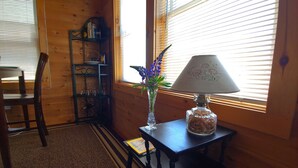 A wooden interior with a table, a lamp, a vase with flowers, and a bookshelf. - Beautiful and modern cape style cottage nested along the Jordan Tidal River. (Lamoine)