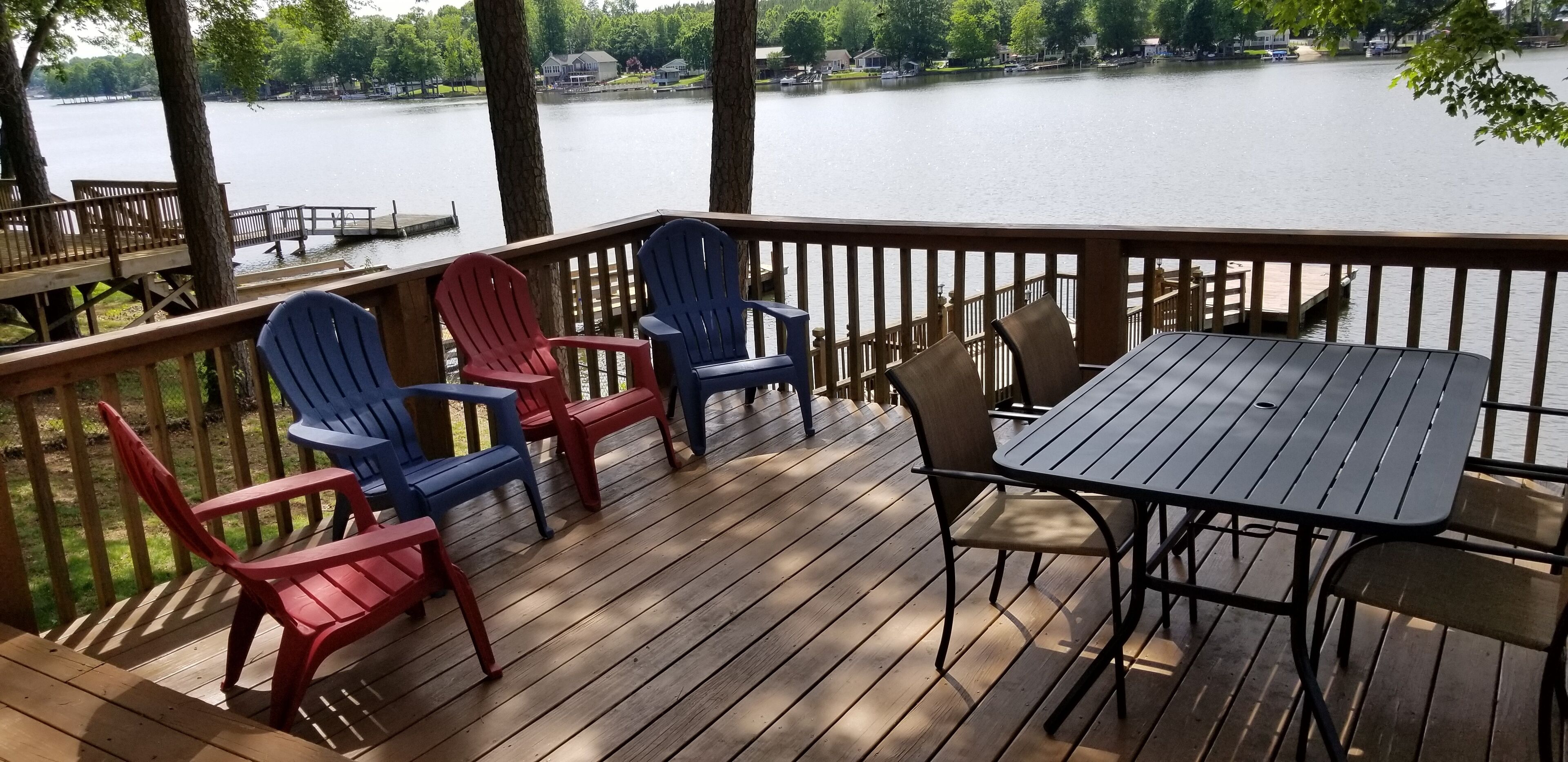 A wooden deck with Adirondack chairs and a table, overlooking a lake with houses in the background.