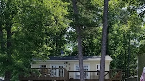 A wooden deck leading to a house with a balcony, surrounded by trees and a body of water. - Lake front cottage with dock (Lexington)