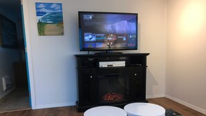 A living room with a flat-screen TV on a black stand, a fireplace, and two white side tables.