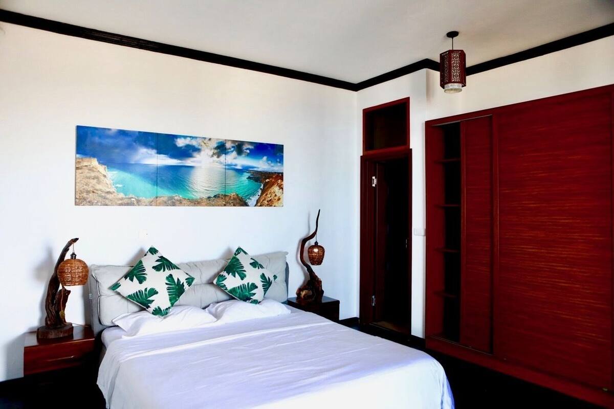 A bedroom with a bed, a nightstand, a wall-mounted painting of a coastal scene, a wooden wardrobe, and a hanging lamp.