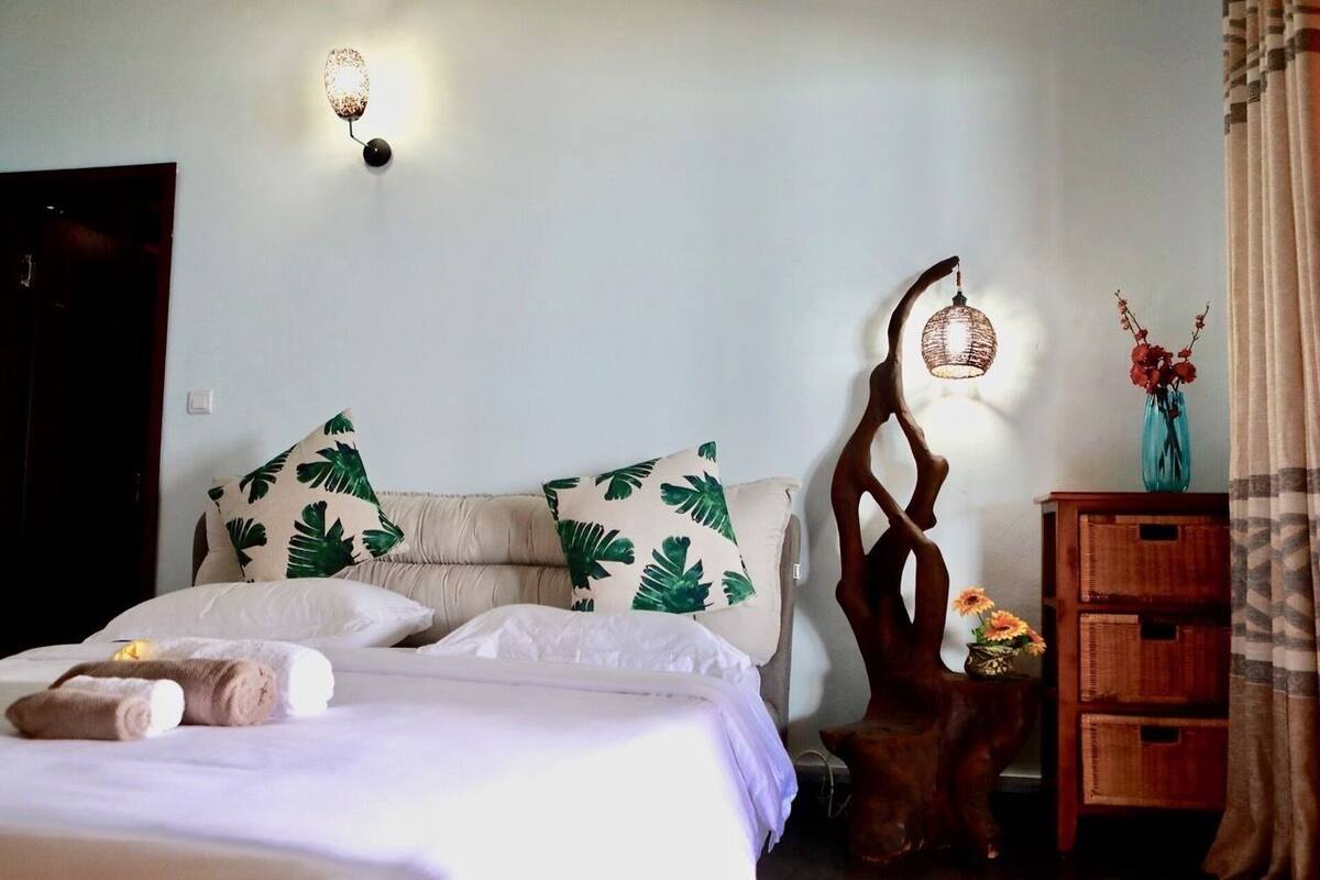 A hotel room with a bed, two pillows with a leaf pattern, a wooden nightstand with a vase of flowers, and a wall-mounted light fixture.