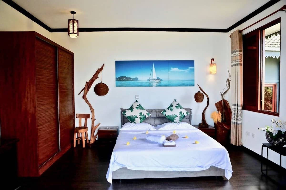 A bedroom with a bed, a nightstand, a wooden wardrobe, a chair, a wall-mounted shelf, and a window with curtains.