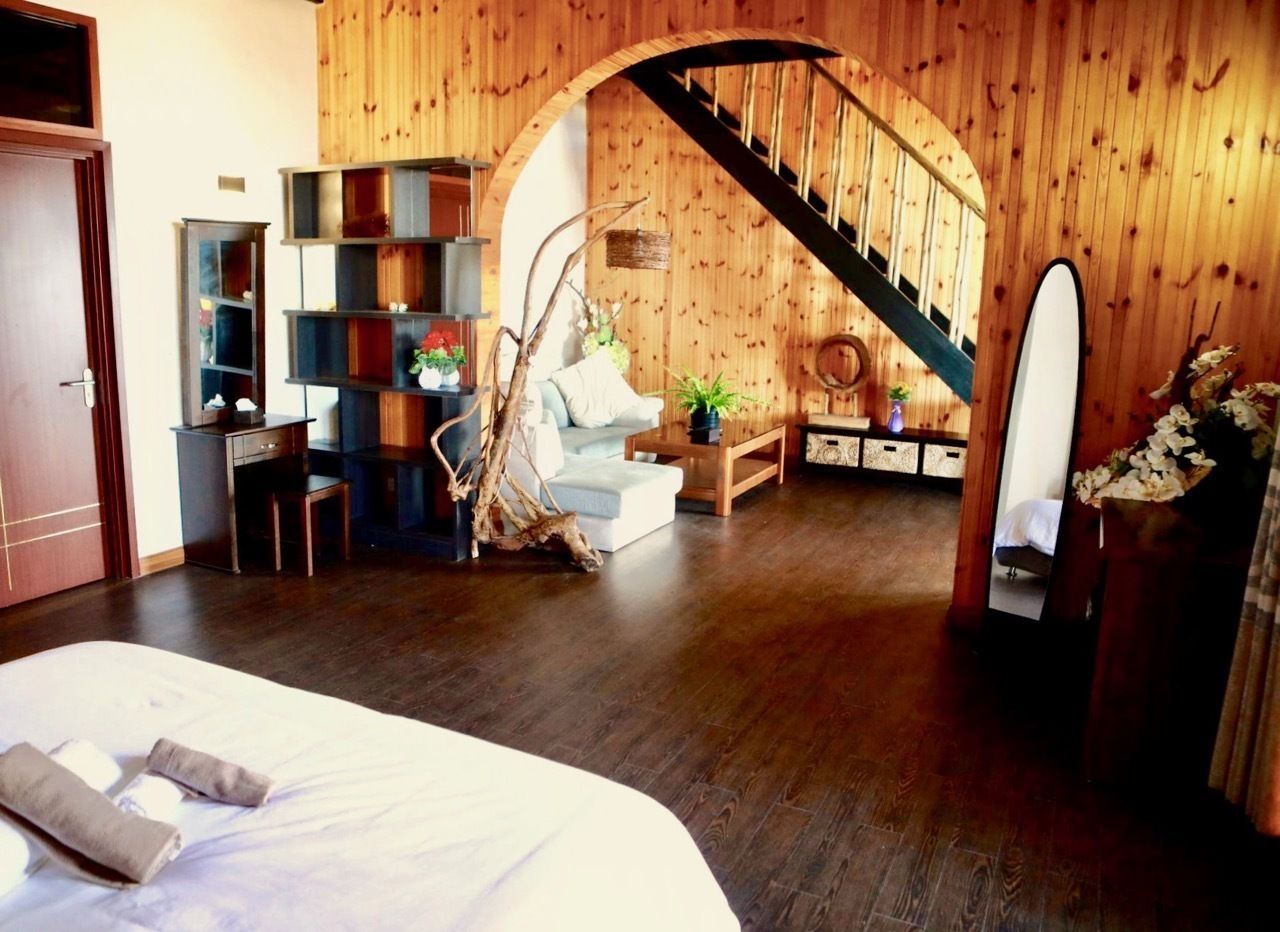 A hotel room with a wooden interior, a bed with white linens, a seating area with a sofa and chairs, a small table with a vase, and a staircase with a wooden railing.