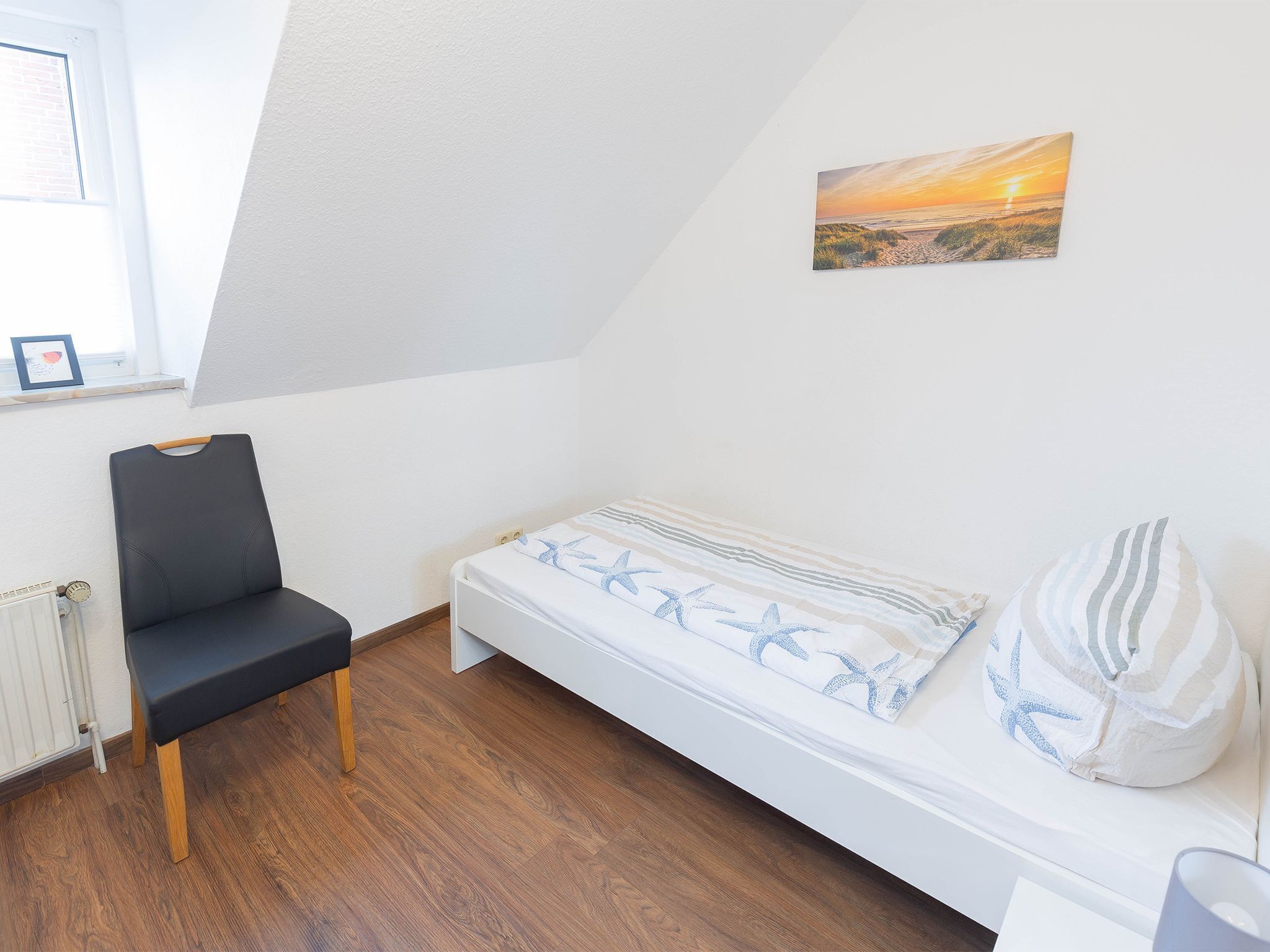 A small, single-room bedroom with a white bed, a black chair, and a framed picture on the wall.