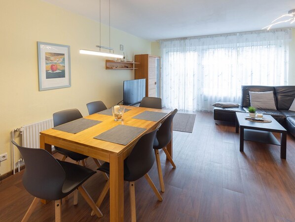 A modern living room with a wooden dining table, black chairs, a leather sofa, and a TV. - Vacation home Marisol, holiday apartment rental North Sea - Holiday home Marisol (Varel)