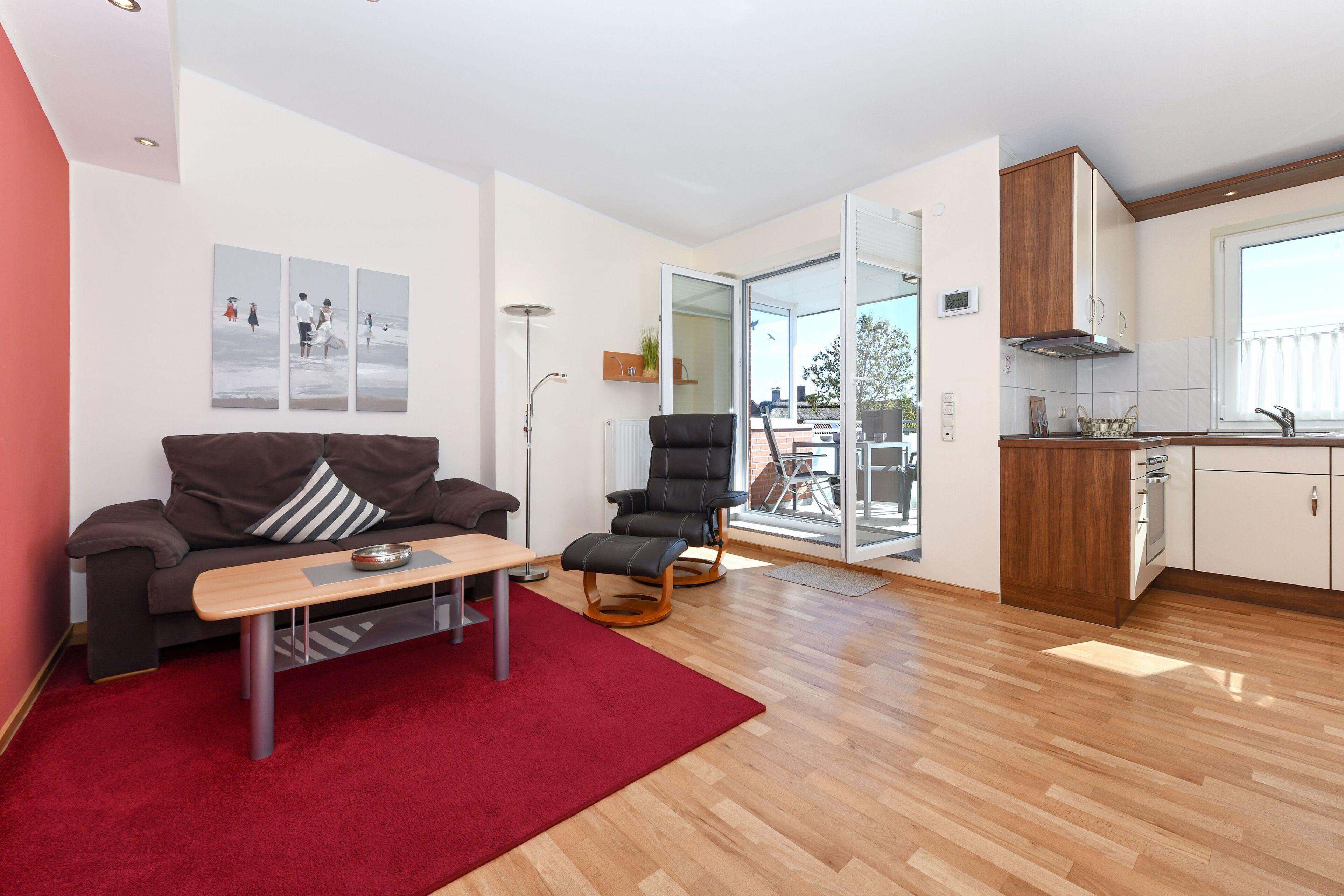 Ein modernes Wohnzimmer mit einer Couch, einem Couchtisch, einem roten Teppich, einem schwarzen Sessel und einer Küchenzeile mit Holzschränken und einem Fenster.