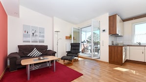 A modern living room with a sofa, a coffee table, a red rug, a black chair, and a kitchen area with wooden cabinets and a window. - House Pier 18 - Holiday apartment 9 (Esens)